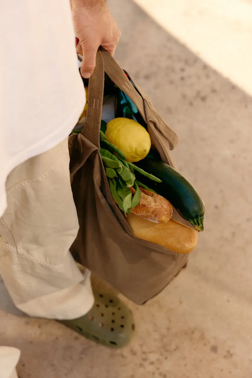 Person carrying a canvas market bag filled with bread, greens, lemon, and zucchini